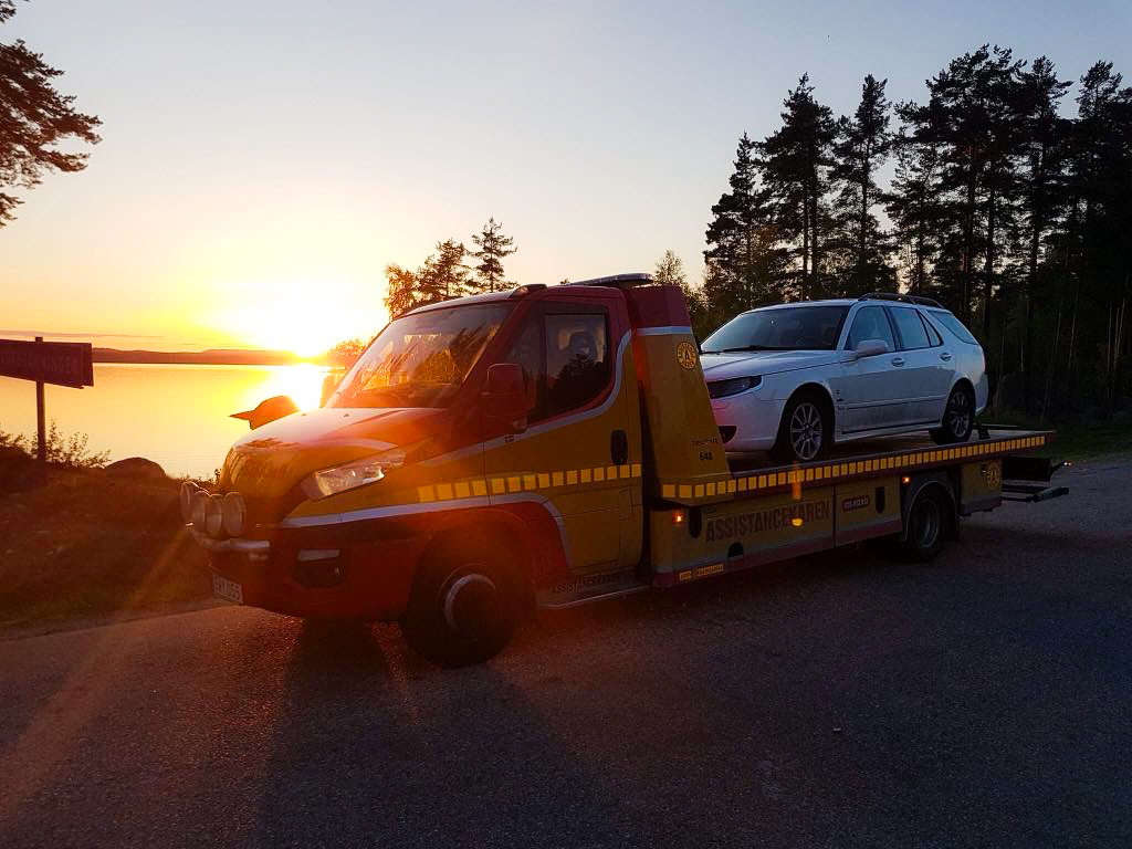 Bärgningsbil med bil på flak vid solnedgång vid sjön, Söderhamns Bärgningstjänst
